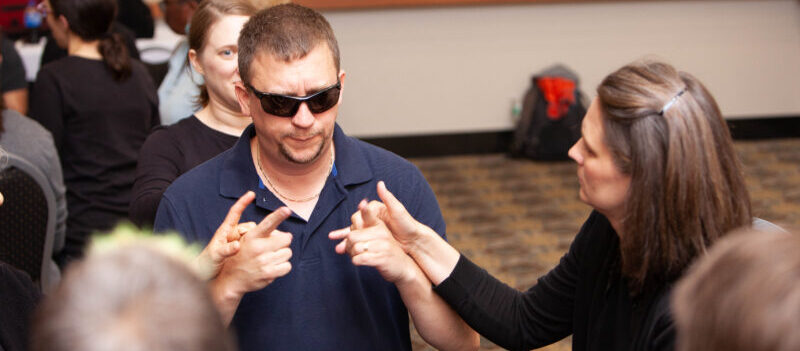A workshop featuring a DeafBlind individual wearing sunglasses, signing using ProTactile ASL, with an interpreter to his right.